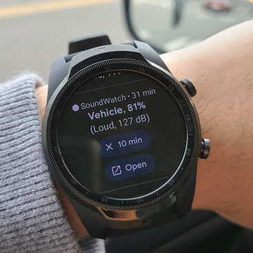 A user is sitting in a car wearing a smartwatch that shows the recognized sound as Vehicle with a confidence level of 81%.